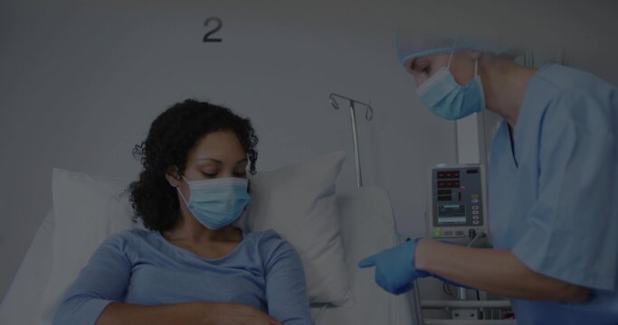 Listening patient in blue top mask on hospital bed, nurse in scrubs gesturing by IV pump