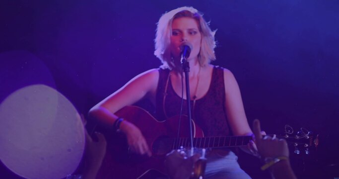 Singing solo performer in sleeveless-top and light pants, playing acoustic guitar into mic on stage