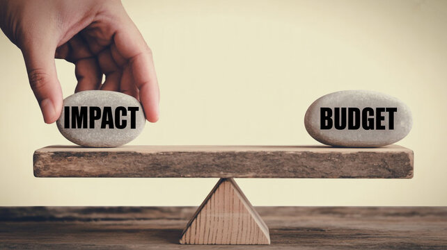 A hand balancing stones with the words impact and budget on a wooden seesaw scale representing a business financial decision or strategy concept for project management and resource allocation