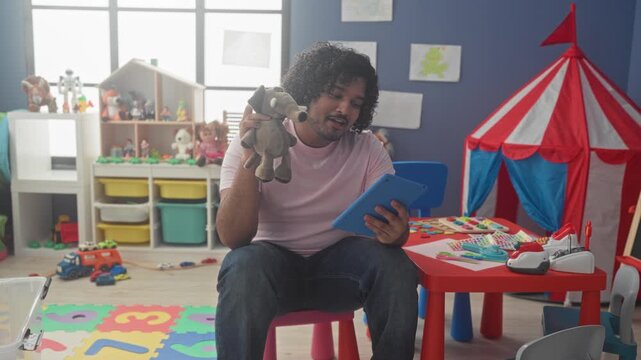 Man with tablet holds plush elephant puppet to face while seated in building playroom; nurturing gentle care.