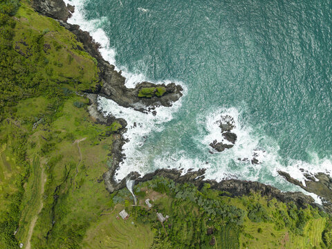 Aerial view of Banyutibo Water Fall cascading into the turquoise ocean with waves crashing against rugged cliffs and lush greenery in Girisubo, Special Region of Yogyakarta, Indonesia.