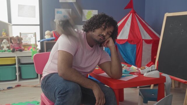 Man holding stuffed elephant, hand raised while sitting at red table with craft toys and circus tent in kindergarten building playroom; pensive.