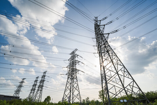 This image shows multiple steel lattice high voltage transmission pylons strung with power lines, set against a bright cloudy daytime sky with green vegetation at the base.