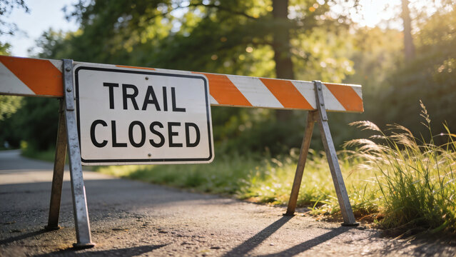 Cinematic eye-level shot of a TRAIL CLOSED sign on an orange and white striped barricade blocking an asphalt path