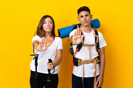 Young mountaineer couple with a big backpack isolated on yellow background making stop gesture