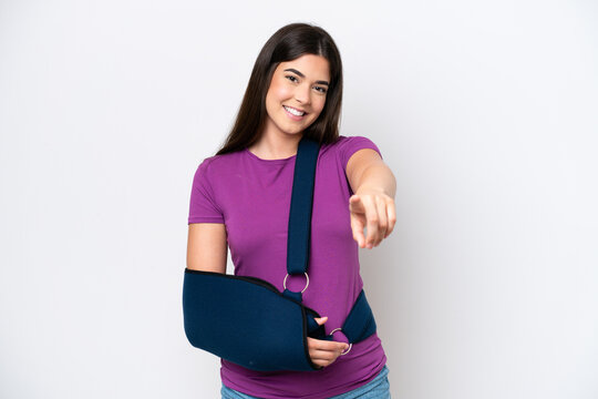 Young Brazilian woman with broken arm and wearing a sling isolated on white background pointing front with happy expression