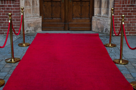 Red carpet pathway with gold stanchions and red velvet ropes leading to a large historical wooden double door entryway of a brick building.