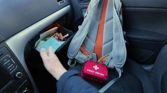 Glovebox First Aid Kit Retrieval From Dashboard Shows Hand Reaching Into Glovebox To Grab Red Pouch, Pulls Out Bandage And Wipes, Stows Items Into Backpack On Seat, Urgent Readiness Mood While Parked.