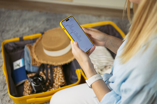 Woman checking travel checklist on smartphone, preparing suitcase for summer vacation, packing holiday essentials, organizing luggage before departure