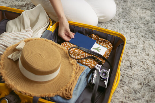 Woman packing yellow suitcase for summer vacation, preparing travel essentials like passport, boarding pass, straw hat, and camera for an upcoming trip abroad