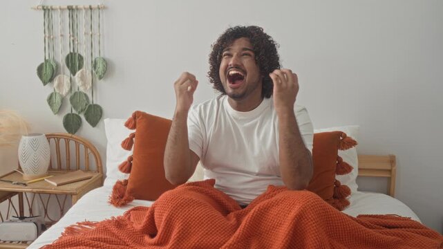 Man on bed with hands on temples screaming for headache in studio with orange blanket and tassel pillows; frustration anxiety.
