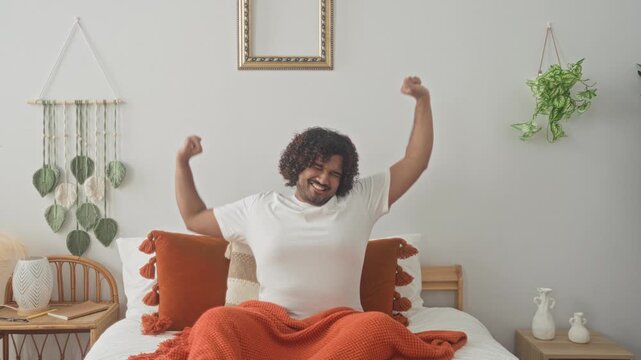 Man on bed stretches arms upward in studio with orange blanket and tassel pillows, hands raised above head in waking stretch; calm morning serenity.