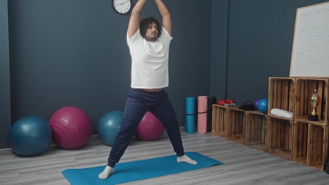 Young man on blue yoga mat stretching with arms raised in studio gym with exercise balls and wooden crates; serenity wellness balance.