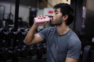 Fototapeta premium Gym fitness man athlete workout hydration muscular man drinking from pink water bottle gym weight room after strength training, wearing gray shirt, showing focus, recovery, wellness, and endurance