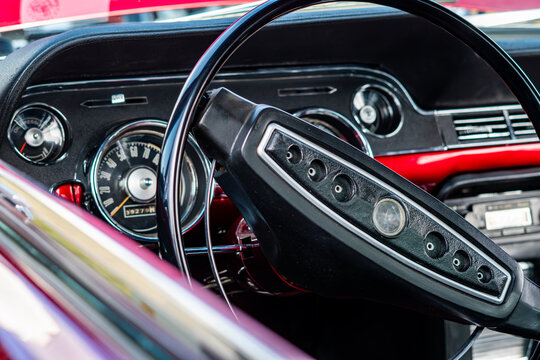 Close-up view of a classic vintage car interior featuring a detailed black steering wheel, dashboard gauges, speedometer, oil pressure meter, and bright red car body in the background, automotive phot