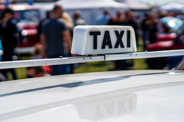 A close-up shot of a classic white vintage "TAXI" roof sign with black lettering mounted on a car, featuring a clear reflection of the text on the vehicle's roof, outdoors, blurred background. © Adam