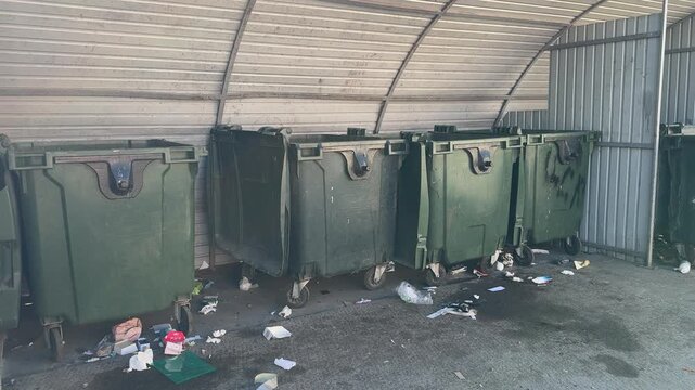 Sunlit covered dumpster area with garbage. Three green bins under metal canopy, litter scattered across stained asphalt, plastic