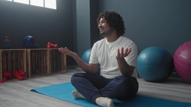 Man seated crosslegged on blue mat gesturing with hands near exercise balls and wooden crates in studio with window light; calm reflection.