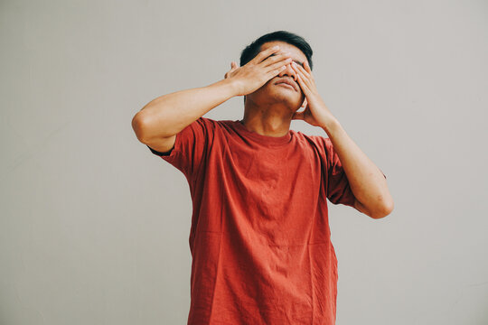 man covering face with hands in red shirt on gray background