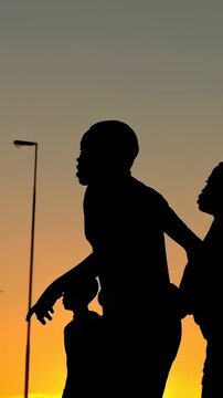 Vertical video: Walking teen wearing tee and shorts leading two boys after ball toss at sunset lamp