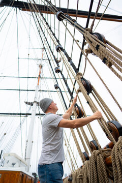 A man works on tying ropes on a tall ship in a harbor. The setting shows masts and rigging with a cloudy sky above. This scene displays the activity of sailing preparation.