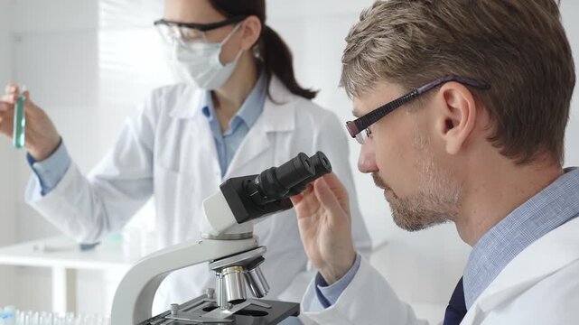 Scientific professional examining specimen through microscope, laboratory assistant nearby wearing protective mask, both dressed in white lab coats during research process. Medicine and science