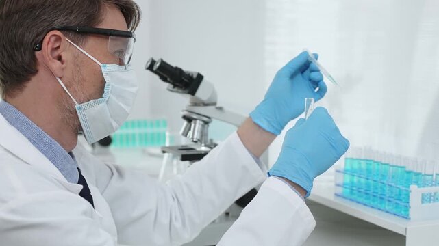 Masked male scientist in lab coat, protective glasses and blue gloves is adding reagent to test tube with microscopes and test tube rack in modern laboratory. Medicine, healthcare and science concept