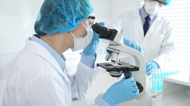 Female scientist wearing protective gear is examining specimen under microscope while man laboratory assistant handles glassware in a professional medical investigation. Medicine and science concept