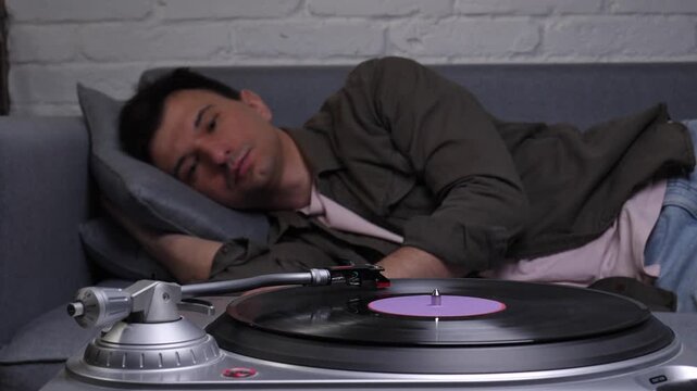 Young man relaxing on a comfortable sofa, intently listening to vintage vinyl music playing on a record player, creating a calm and nostalgic atmosphere at home