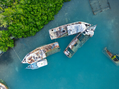 Aerial view of several rusty shipwrecks abandoned in turquoise water near a lush green mangrove shoreline Guanaja, Bay Islands Department, Honduras.