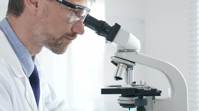 Adult male scientific researcher analyzing sample through microscope, wearing protective gear in high tech laboratory environment. Healthcare, medicine and science concept