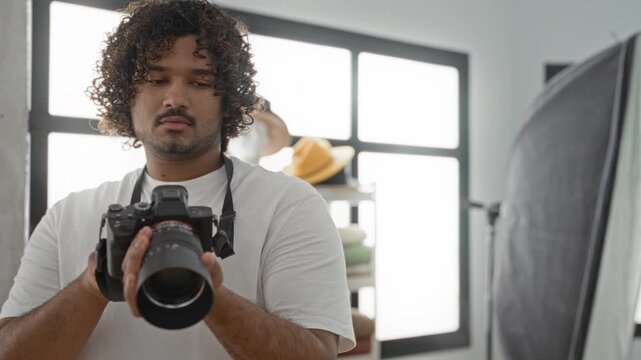 Man holding camera with hands and adjusting lens in studio near softbox and hat shelf, checking settings on lcd; quiet concentration.
