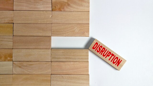 A wooden block with the word "DISRUPTION" in red text is pulled out from an organized wall of plain blocks, illustrating a break in structure or a shift in a standard process against a white surface.