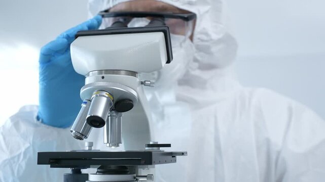 Medical virologist in a hazmat suit, mask, and blue gloves, carefully working at a microscope in a laboratory researching viruses. Healthcare, medicine and science concept