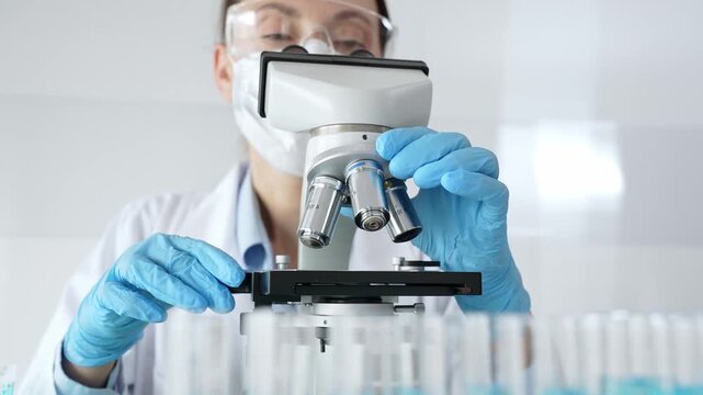 Female scientist wearing protective equipment checking microscope, analyzing blue test tubes in modern laboratory research setting. Medicine, healthcare and science concept