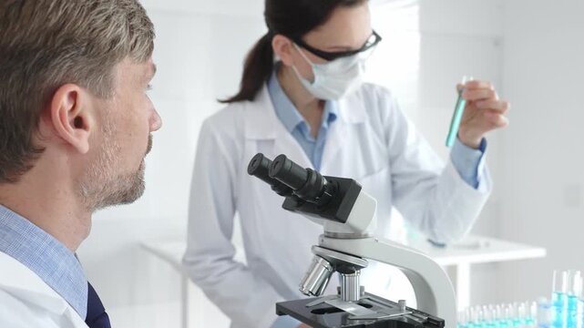 Scientific professional examining specimen through microscope, laboratory assistant nearby wearing protective mask, both dressed in white lab coats during research process. Medicine and science