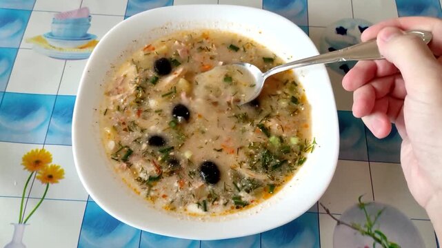 Person stirring solyanka soup with olives and sausage in bowl, first person view, home kitchen setting