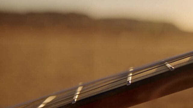 Slow motion tracking pan of guitar neck strings being strummed and sliding across frets in desert dunes at golden sunset evening