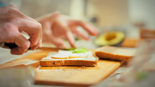 In a home kitchen, a chef's skilled hands prepare a nutritious breakfast by making toast with ham and fresh green avocado, embodying the healthy breakfast concept with a delicious meal