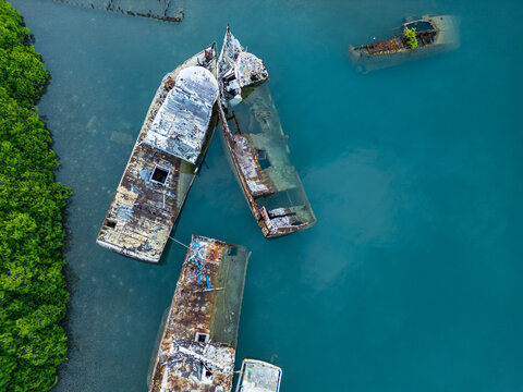 Aerial view of rusty shipwrecks and sunken boats in turquoise water near lush green mangroves in Guanaja, Bay Islands Department, Honduras.