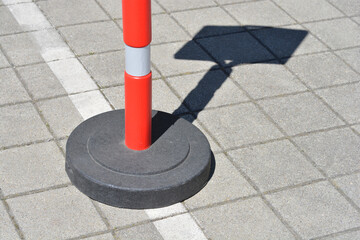 A red and white plastic traffic delineator post with a heavy black base stands on a grey paved surface casting a dark shadow © nahhan