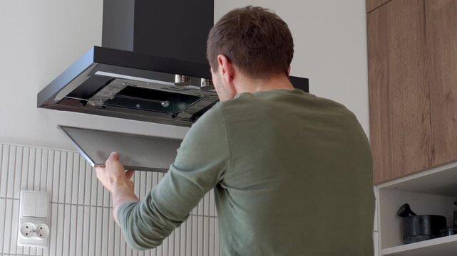 Rear view of a man removing the metal filter from a modern black kitchen hood during cleaning or maintenance in a home kitchen.