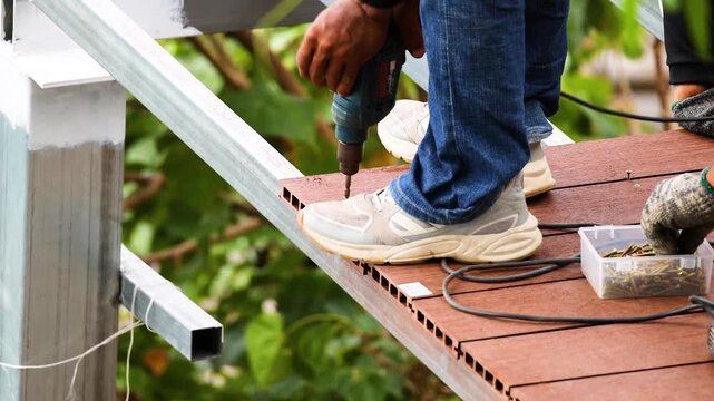 Construction worker using a drill to attach wooden planks to metal frame for deck building.