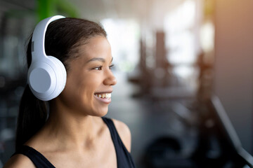 Side profile portrait of smiling young Black woman in gym wearing white wireless headphones, black tank top, happy expression looking away, blurred gym equipment in background © Anastasiya