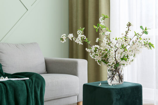 Interior of light living room with blossoming branches in vase, sofa and pouf