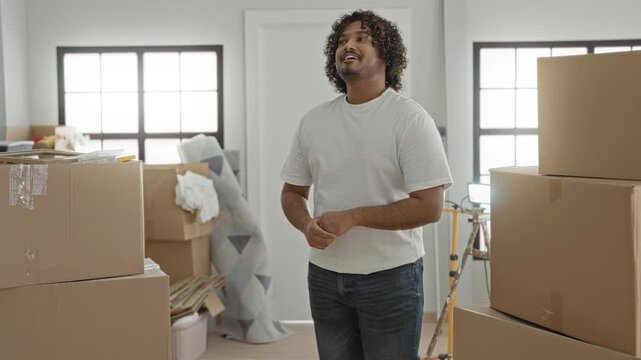 Young indian man smiling with arms crossed beside stacked cardboard moving boxes in building during moving in; contentment.