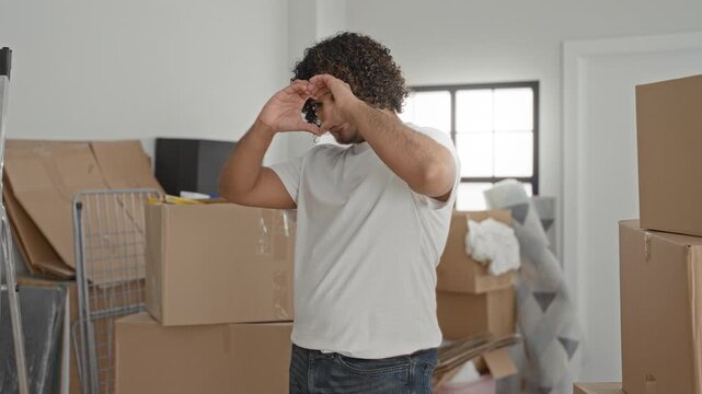 Young indian man wearing white t shirt forms heart with hands in a building amid stacked moving boxes, mattress and packing supplies while smiling and unpacking; home love joy.
