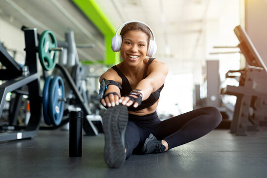 Smiling young Black woman sitting on gym floor stretching legs forward, wearing white headphones, fingerless gloves and phone armband, water bottle beside her, bright gym equipment background