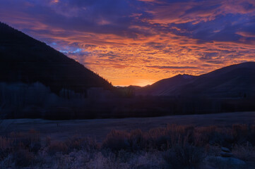 Obraz premium Majestic mountains frame a fiery sunset sky. Valley floor glows with soft, silvery frost. Deep purples blend with orange hues in clouds. Silent, frost-kissed grasses dot the foreground