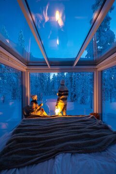 Two travelers enjoy a serene evening in a glass cabin in Akaslompolo, surrounded by pristine snow-covered trees. The calming ambiance evokes a sense of tranquility and adventure in nature.